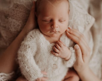 May include: A newborn baby wearing a soft, white, button-up onesie. The baby is nestled in someone's arms, with closed eyes. The onesie has long sleeves and a fuzzy texture. The overall image has a soft, warm tone.