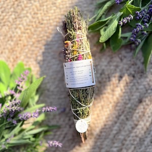 Könnte beinhalten: Ein Bündel getrockneter Kräuter, darunter Lavendel und Salbei, umwickelt mit weißer Schnur. Ein rechteckiges Papieretikett mit Text ist in der Mitte befestigt, und ein kleines rundes weißes Schild hängt unten. Frische Lavendelzweige liegen daneben.