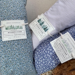Könnte beinhalten: Drei rechteckige Augenkissen mit Stoffbezügen in blauem Blumenmuster, weiß-grauen Streifen und dunkelblauem Blattmuster. Jedes Kissen hat ein weißes Papieretikett, das mit einer weißen Kordel befestigt ist und die Aufschrift Minoa Handmade, Augenkissen trägt.