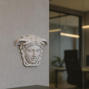 Könnte beinhalten: Eine weiße, geformte Wanddekoration, die ein klassisches weibliches Gesicht mit detaillierten Haaren und Hörnern darstellt. Die Skulptur ist an einer hellgrauen Wand angebracht, mit einer dunklen Holzoberfläche im Vordergrund.