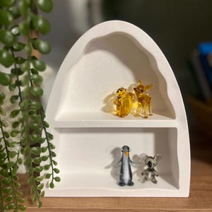 May include: A white, arched shelf unit displays four small glass animal figurines. The top shelf holds a yellow squirrel and a deer. The bottom shelf features a penguin and a koala bear. Green leafy plant in the background.