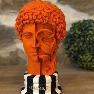 Puede incluir: Escultura naranja de un busto clásico con un peinado rizado y un diseño dividido. La base es a rayas blancas y negras con la palabra "HERMES" en letras doradas. El busto es un objeto decorativo.