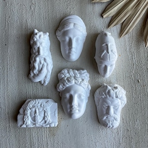 Könnte beinhalten: Eine Sammlung von sechs weißen Gips-Skulpturen klassischer Büsten und Gesichter. Die Skulpturen zeigen verschiedene Figuren mit detaillierten Gesichtszügen, darunter lockiges Haar, Bärte und Helme. Die Büsten sind auf einer strukturierten, hellfarbigen Oberfläche angeordnet.