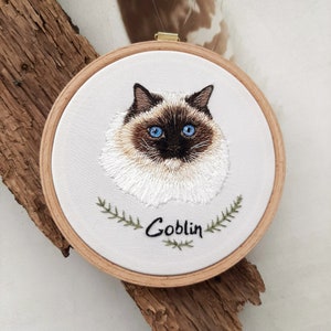 Op de afbeelding: Borduurwerk van een wit kat met blauwe ogen in een ronde houten ring. De naam van de kat, "Goblin", is geborduurd onder de afbeelding van de kat.