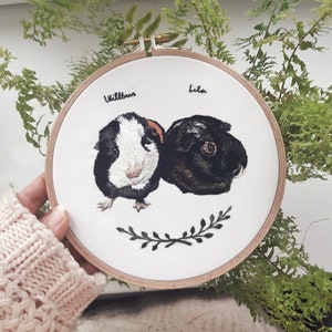 Könnte beinhalten: Stickerei auf einem runden Rahmen mit zwei Meerschweinchen, eines schwarz-weiß und eines schwarz, mit ihren Namen "Wilbur" und "Lila" darüber gestickt. Unter den Meerschweinchen befindet sich ein schwarzes gesticktes Blumenmuster.