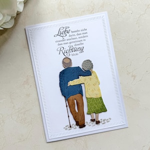 Op de afbeelding: Een witte wenskaart met een uitgesneden illustratie van een ouder echtpaar dat samen loopt. De persoon links draagt een blauw jasje en bruine broek met een wandelstok, terwijl de persoon rechts een geel topje en groene rok draagt. De tekst luidt: Liebe besteht nicht darin, dass man einander anschaut, sondern dass man gemeinsam in dieselbe Richtung blickt.