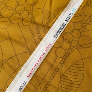 Op de afbeelding: Mosterdkleurige stof met een geometrisch patroon van cirkels en lijnen. Een wit label toont de tekst "227558 MARCH BALLOONS FRANK LLOYD W". Geschikt voor kleding of woninginrichting.