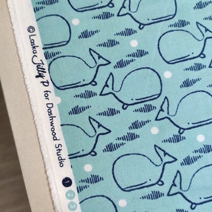 May include: Light blue fabric with a repeating pattern of whales, clouds, and white dots. The whales are outlined in dark blue. The text "Laska Tilly P for Dashwood Studio" is printed on the edge of the fabric.