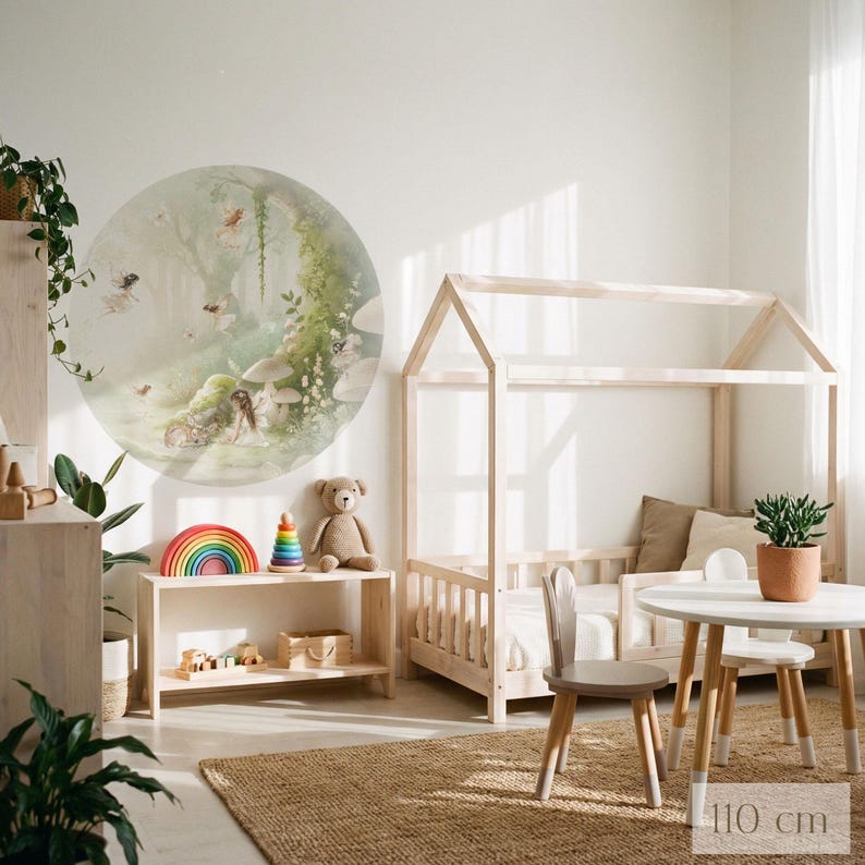 K&ouml;nnte beinhalten: Ein Kinderzimmer-Set aus hellem Holz mit einem hausf&ouml;rmigen Bettrahmen, einem kleinen Tisch und St&uuml;hlen. Ein gro&szlig;er runder Wandaufkleber zeigt Feen in einer Waldszene. Ein Holzregal zeigt Spielzeug, darunter einen Regenbogenstapler.