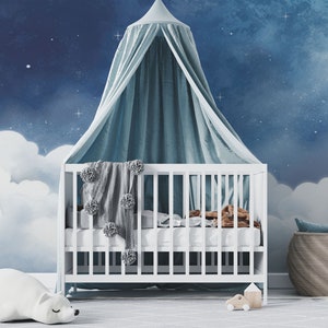 May include: A white crib with a blue and white canopy, a white stuffed animal, and a white blanket with gray pom-poms. The crib is in front of a blue and white wall mural with clouds and stars.