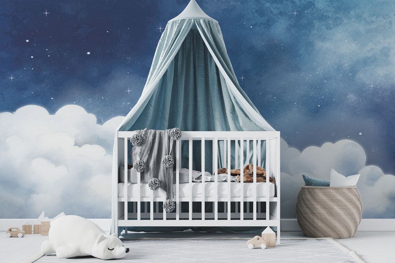 May include: A white crib with a blue and white canopy, a white stuffed animal, and a white blanket with gray pom-poms. The crib is in front of a blue and white wall mural with clouds and stars.