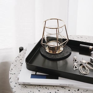 Könnte beinhalten: Ein schwarzes Tablett mit einem goldenen geometrischen Kerzenhalter, einem grauen Filzuntersetzer und goldenem Schmuck auf einer weiß-grauen, gesprenkelten Oberfläche. Das Tablett liegt auf einem Buch mit dem sichtbaren Text "YVES KLEIN" und "TASCHEN".