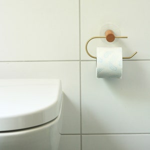 May include: A white toilet paper roll with a blue feather pattern is on a gold metal toilet paper holder with a wooden base. The holder is mounted on a white tiled wall.
