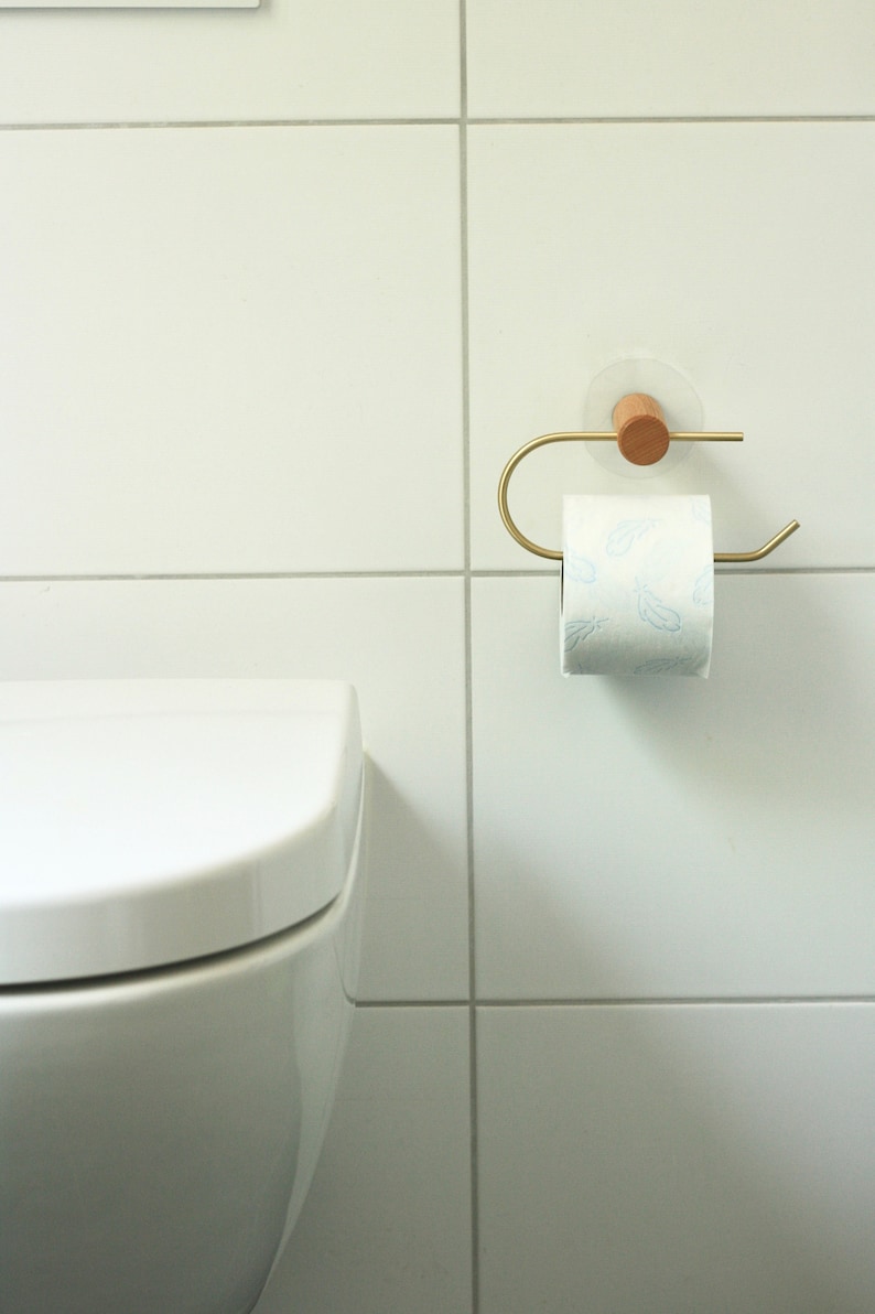 May include: A white toilet paper roll with a blue feather pattern is on a gold metal toilet paper holder with a wooden base. The holder is mounted on a white tiled wall.