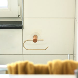 May include: A gold metal paper towel holder with a wooden knob attached to a white tiled wall.