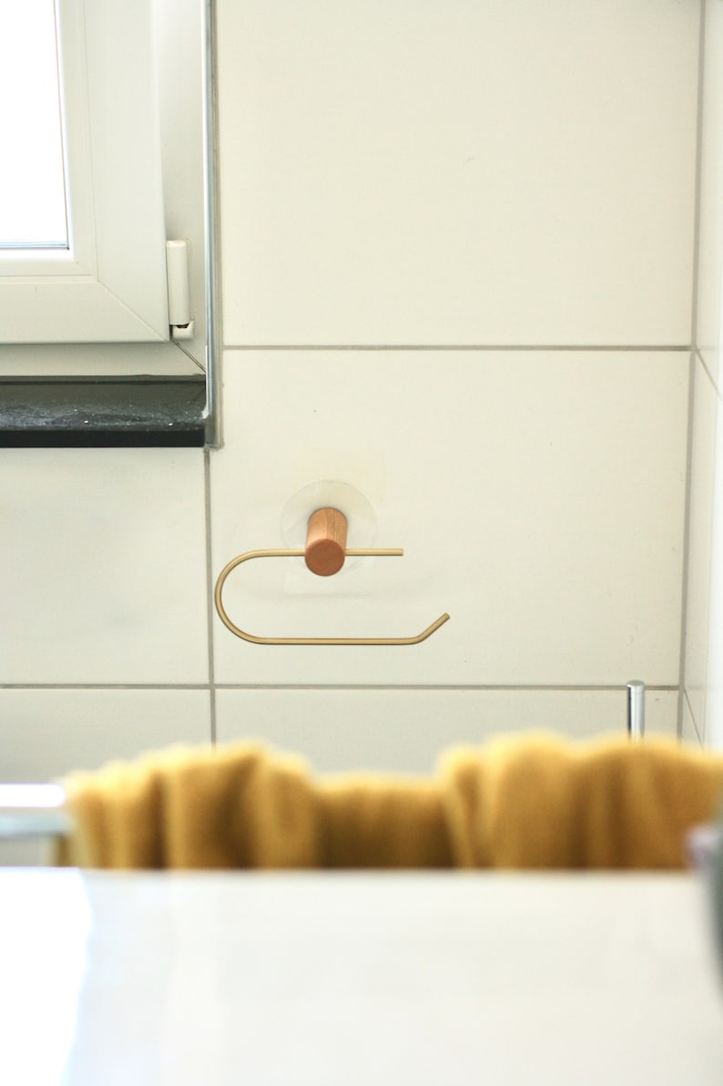 May include: A gold metal paper towel holder with a wooden knob attached to a white tiled wall.