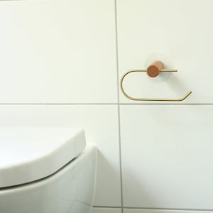 May include: A gold metal toilet paper holder with a wooden base mounted on a white tiled wall. The holder is attached to the wall with a white adhesive disc.