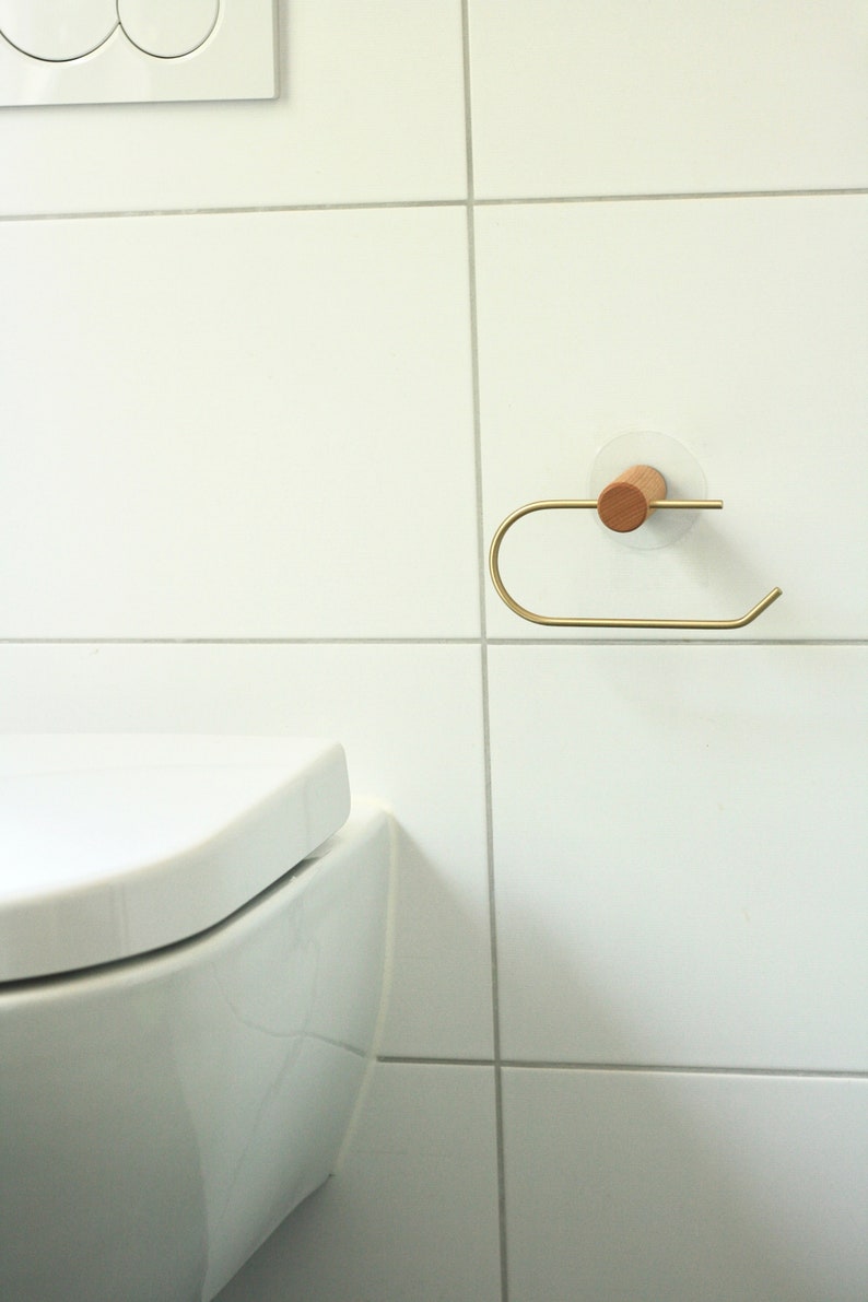 May include: A gold metal toilet paper holder with a wooden base mounted on a white tiled wall. The holder is attached to the wall with a white adhesive disc.