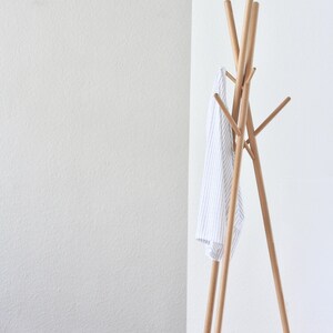 May include: A wooden coat rack with a minimalist design. The rack has a tripod base and multiple arms for hanging clothes. A white and blue striped shirt hangs from the rack. The rack is made of light-colored wood.