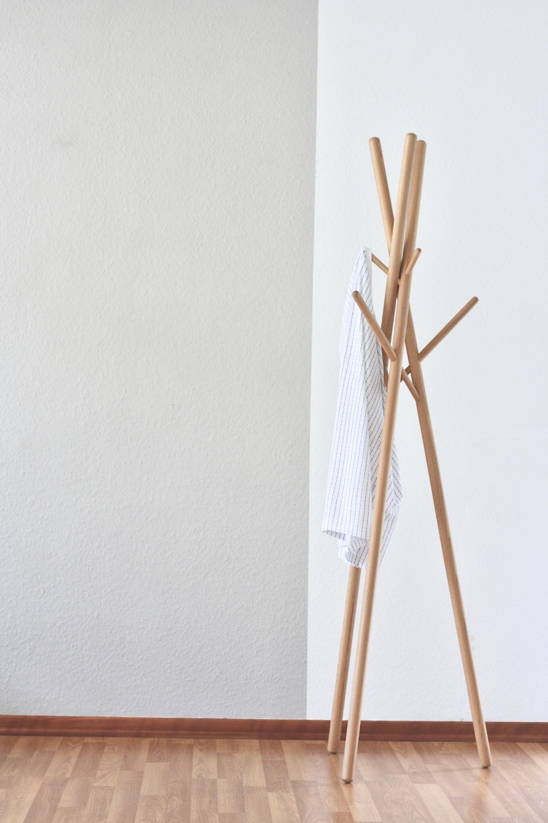 May include: A wooden coat rack with a minimalist design. The rack has a tripod base and multiple arms for hanging clothes. A white and blue striped shirt hangs from the rack. The rack is made of light-colored wood.