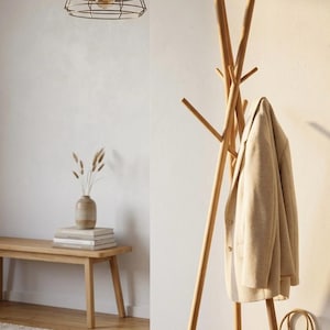 May include: A wooden coat rack with a beige jacket hanging on it. A woven basket and white sneakers sit on the floor. A bench with books and a vase with dried plants are also visible.