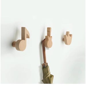 Könnte beinhalten: Drei Wandhaken aus Holz in Form einer Musiknote, einer Gitarre und einer Handgeste. Die Haken sind aus hellfarbigem Holz gefertigt und an einer weißen Wand montiert. Ein Regenschirm hängt am Gitarrenhaken.