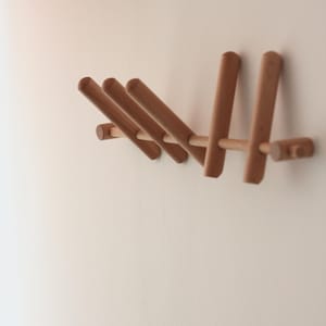 Op de afbeelding: Een houten wandkapstok met een minimalistisch ontwerp. De kapstok heeft verschillende cilindrische pinnen die uit een horizontale balk steken. Het hout heeft een natuurlijke, lichtbruine kleur en de kapstok is op een witte muur gemonteerd.