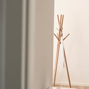 May include: A wooden coat rack with multiple arms and a tripod base. A white umbrella hangs from one of the arms. The rack is made of light-colored wood and stands against a white wall.