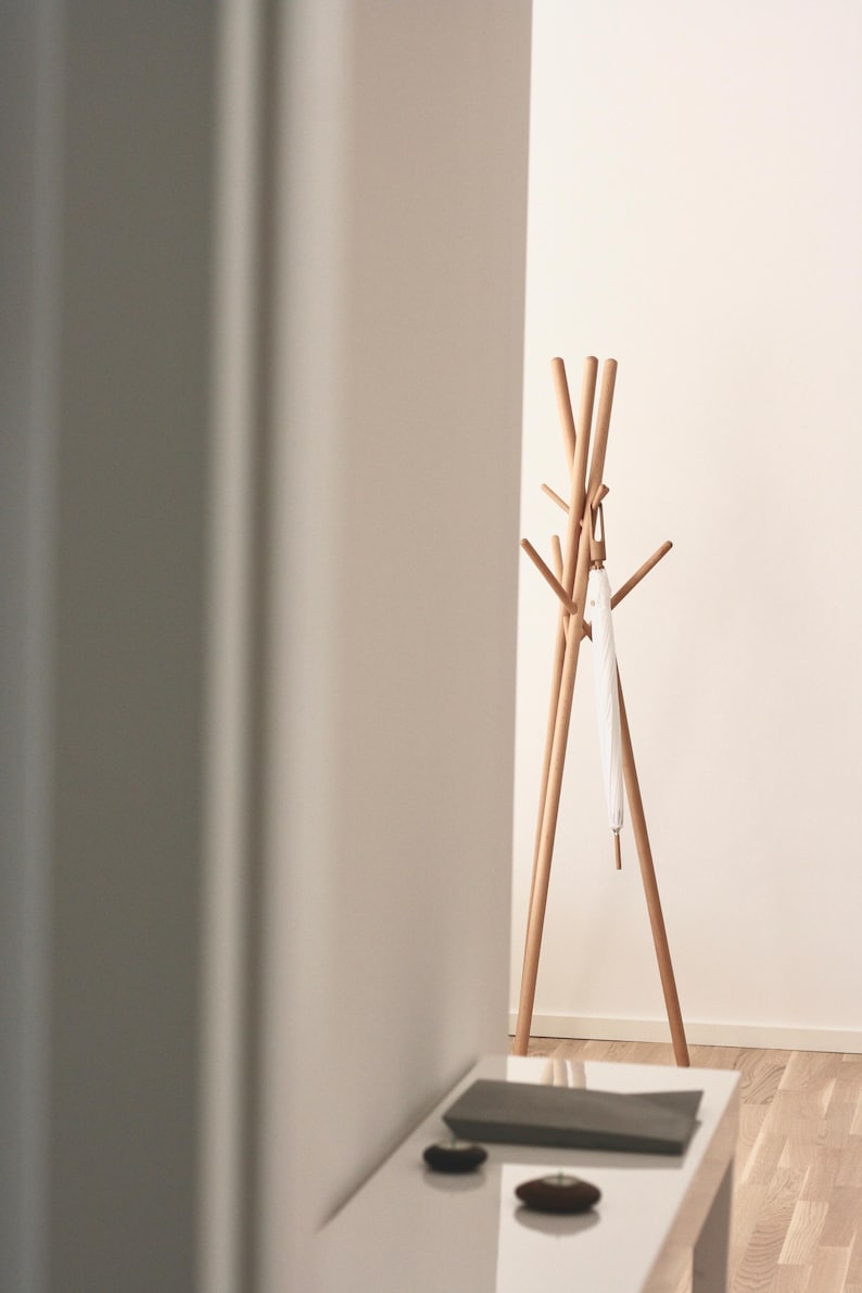 May include: A wooden coat rack with multiple arms and a tripod base. A white umbrella hangs from one of the arms. The rack is made of light-colored wood and stands against a white wall.