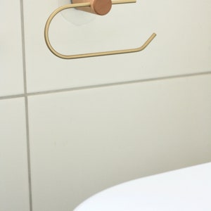 May include: A gold metal toilet paper holder with a wooden base. The holder is mounted to a white tiled wall.