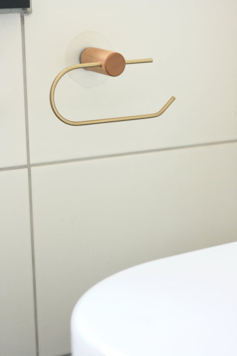 May include: A gold metal toilet paper holder with a wooden base. The holder is mounted to a white tiled wall.