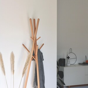 May include: A wooden coat rack with multiple arms, holding a gray scarf. The rack is made of light-colored wood and stands against a white wall. Dried ornamental grass adds a natural touch to the decor.