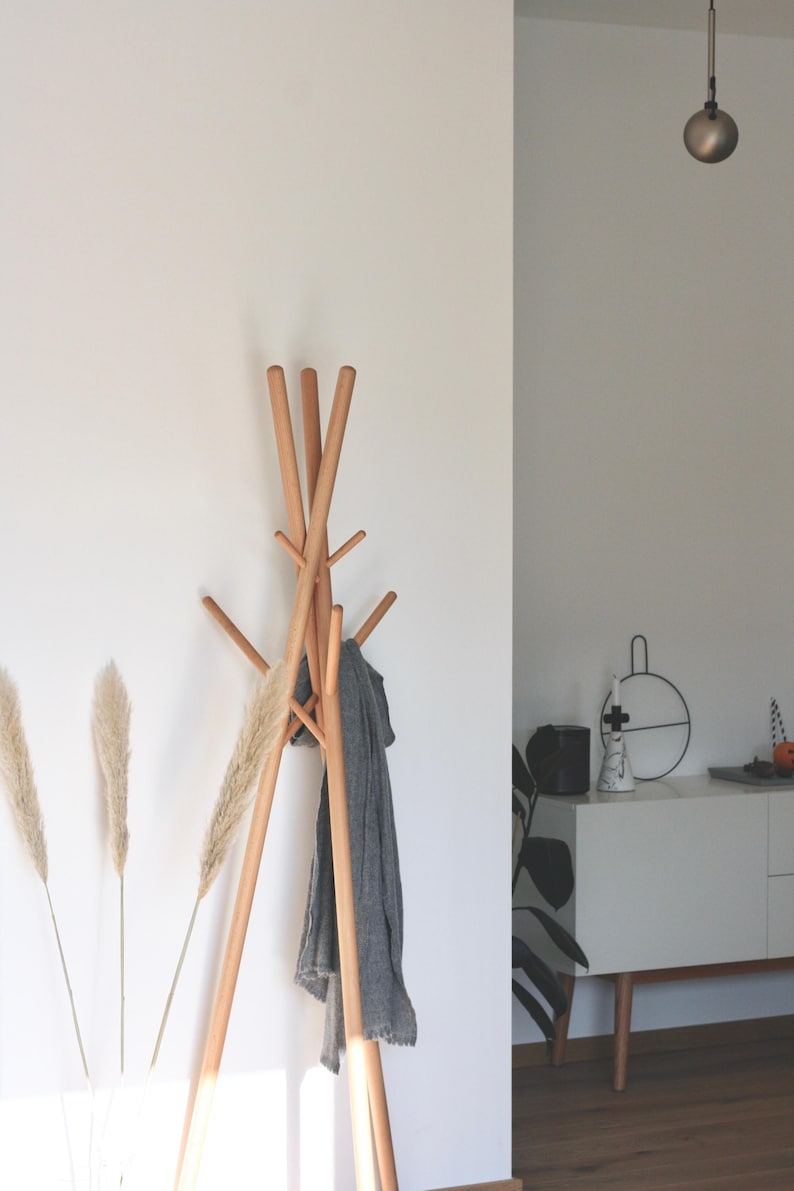 May include: A wooden coat rack with multiple arms, holding a gray scarf. The rack is made of light-colored wood and stands against a white wall. Dried ornamental grass adds a natural touch to the decor.