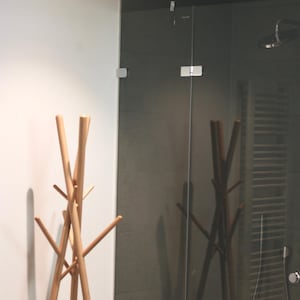 May include: A light-colored wooden coat rack with multiple arms stands in front of a dark gray tiled wall and a glass shower enclosure. The rack's design is minimalist and modern, with a natural wood finish.