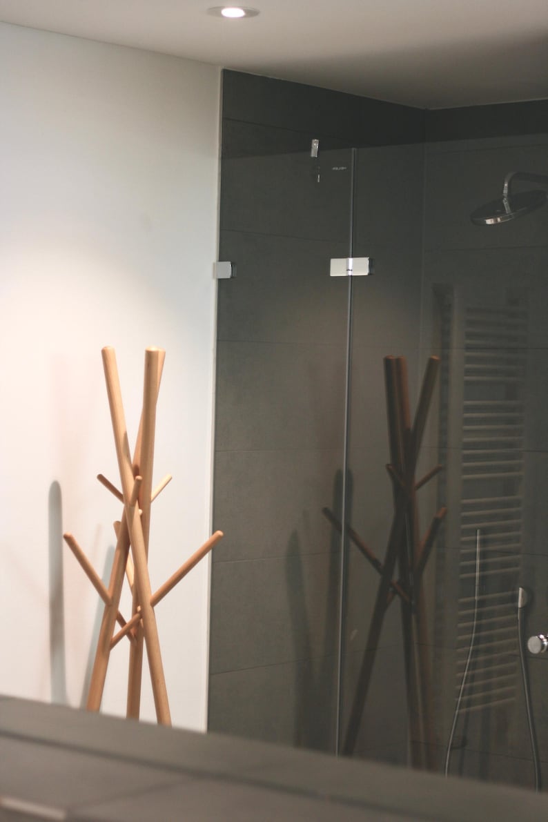 May include: A light-colored wooden coat rack with multiple arms stands in front of a dark gray tiled wall and a glass shower enclosure. The rack's design is minimalist and modern, with a natural wood finish.