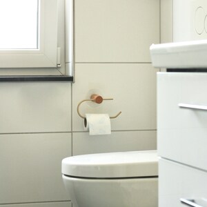 May include: A white toilet with a white toilet paper roll on a gold toilet paper holder with a wooden knob. The toilet is in a white tiled bathroom.