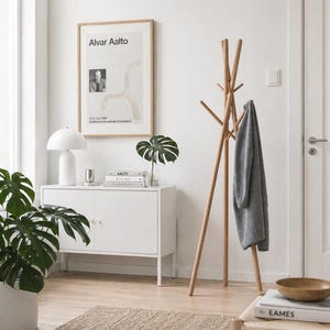 May include: A bright, airy interior scene featuring a wooden coat rack with a gray garment, a white cabinet, and a framed poster. A potted plant and a woven rug add texture. The poster reads "Alvar Aalto".