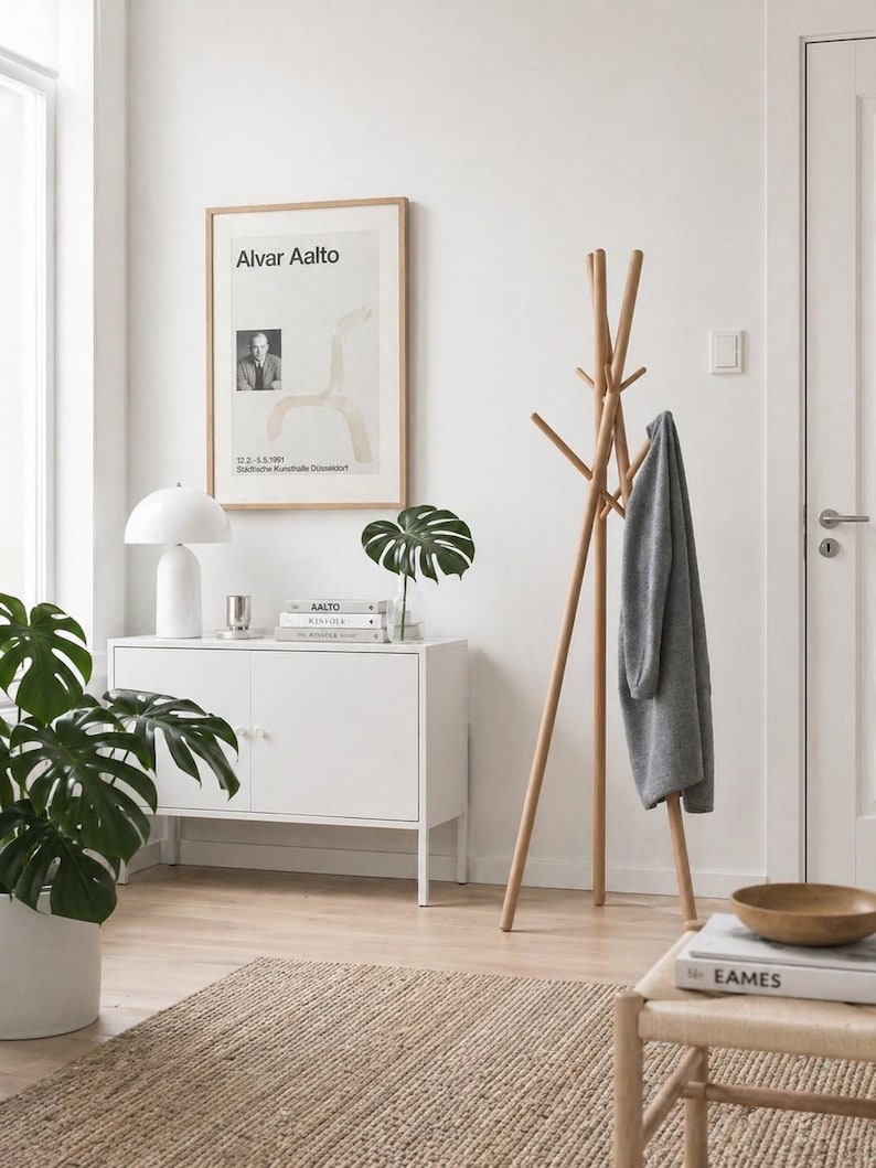 May include: A bright, airy interior scene featuring a wooden coat rack with a gray garment, a white cabinet, and a framed poster. A potted plant and a woven rug add texture. The poster reads "Alvar Aalto".