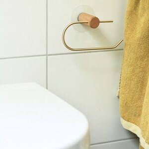 May include: A gold-colored toilet paper holder with a wooden knob is mounted on a white tiled wall. A yellow towel hangs on the wall to the right of the holder.