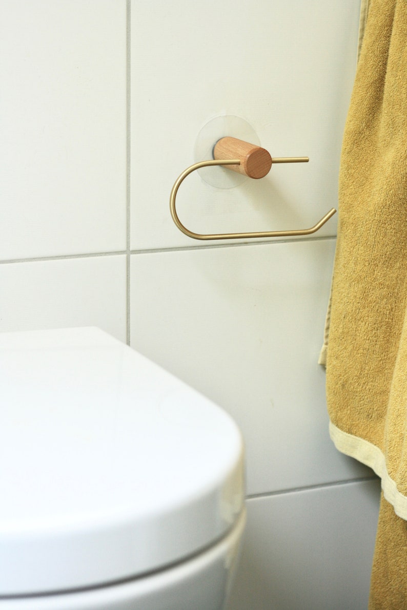 May include: A gold-colored toilet paper holder with a wooden knob is mounted on a white tiled wall. A yellow towel hangs on the wall to the right of the holder.