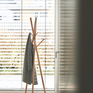 May include: A wooden coat rack with a tripod base and multiple arms for hanging garments. A gray coat hangs on the rack. The rack is positioned in front of a window with horizontal blinds, with natural light illuminating the scene.