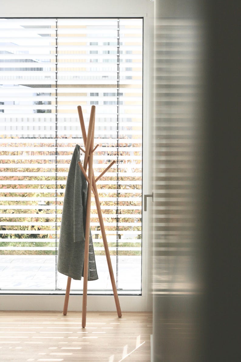 May include: A wooden coat rack with a tripod base and multiple arms for hanging garments. A gray coat hangs on the rack. The rack is positioned in front of a window with horizontal blinds, with natural light illuminating the scene.