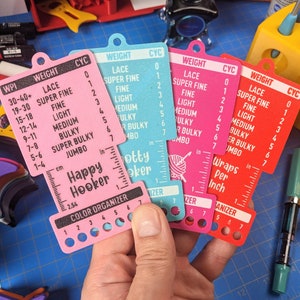 May include: Four colorful plastic gauge cards with a yarn weight and stitch count guide. The cards are labeled "Lace", "Super Fine", "Fine", "Light", "Medium", "Bulky", "Super Bulky", and "Jumbo". The cards also have a ruler in inches and centimeters. The cards are labeled "Happy Hooker" and "Color Organizer".