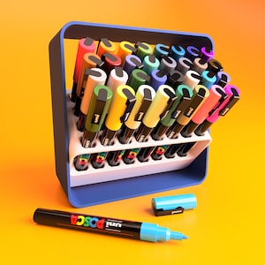 May include: A set of 30 colorful Uni Posca paint markers in a blue and white plastic holder. The markers are arranged in a grid pattern and are all different colors. There are two additional markers lying on a yellow surface.
