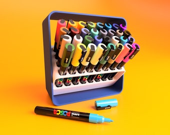 May include: A set of 30 colorful Uni Posca paint markers in a blue and white plastic holder. The markers are arranged in a grid pattern and are all different colors. There are two additional markers lying on a yellow surface.