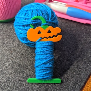 May include: A ball of blue yarn with an orange pumpkin-shaped yarn holder. The yarn holder has a green stem. Other craft supplies are visible in the background.