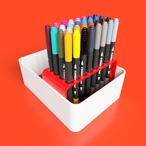 May include: A red plastic organiser holds a collection of colourful markers. The markers are arranged in rows and are all the same size. The organiser is sitting on a white surface.