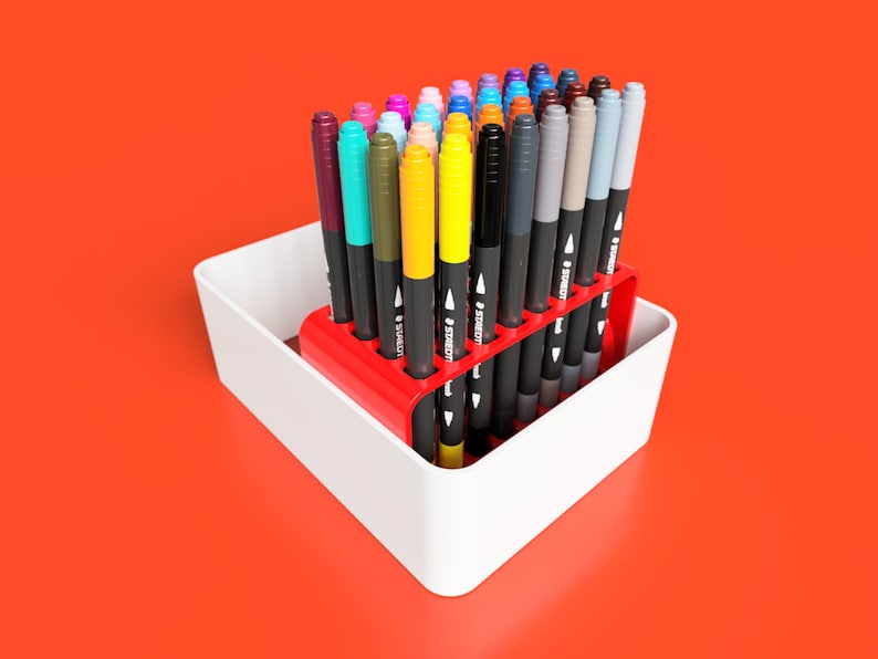 May include: A red plastic organiser holds a collection of colourful markers. The markers are arranged in rows and are all the same size. The organiser is sitting on a white surface.
