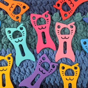 May include: A collection of ten colorful plastic cat-shaped yarn holders. The holders are in various colors, including blue, pink, orange, purple, and yellow. Each holder has a cat face with a small hole in the center for holding yarn.