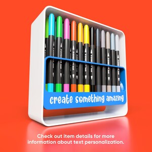 May include: A white holder with a blue section that says "create something amazing" holds a variety of coloured markers.